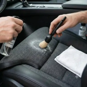 Scrubbing fabric vehicle upholstery gently with a soft brush to demonstrate how to clean car seat safely without damage.